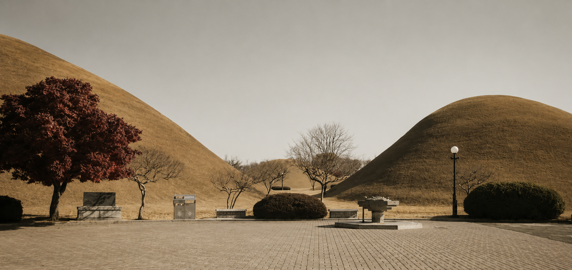 Sepia-toned landscape photograph of a Korean ancestral cemetery with large earth mounds, a paved foreground, a red-leafed tree on the left, and a few bare trees in the distance under a pale sky.